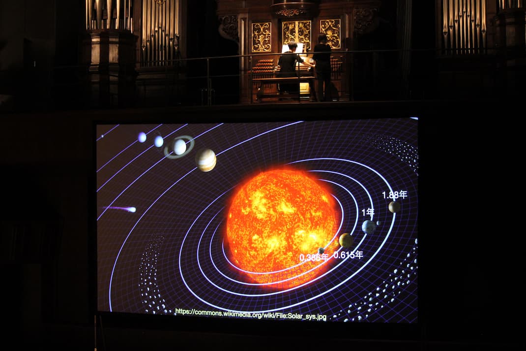 太陽系の大スクリーンを前にパイプオルガンを弾く﨑山